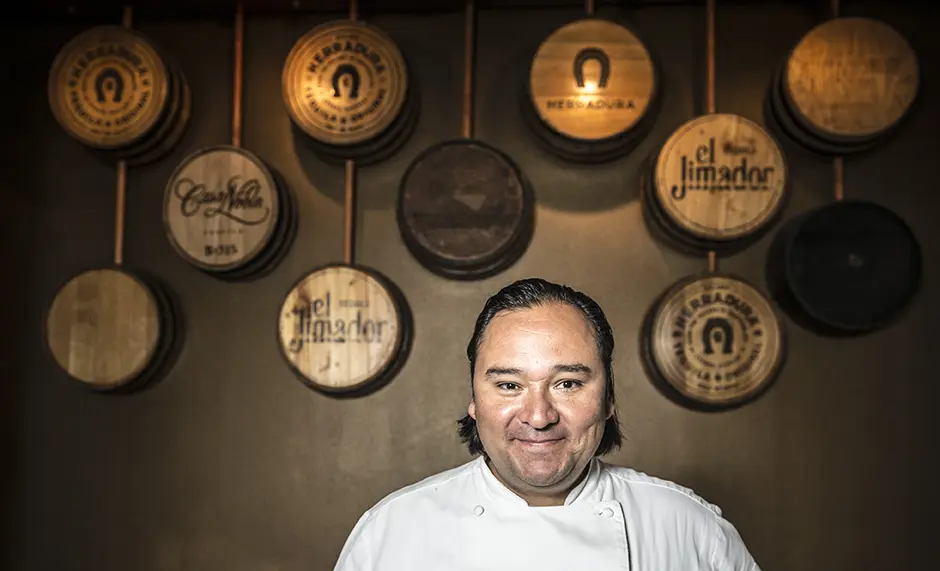 Chef Johnny Hernandez in chef's whites smiling in front of a wall of tequila barrel heads