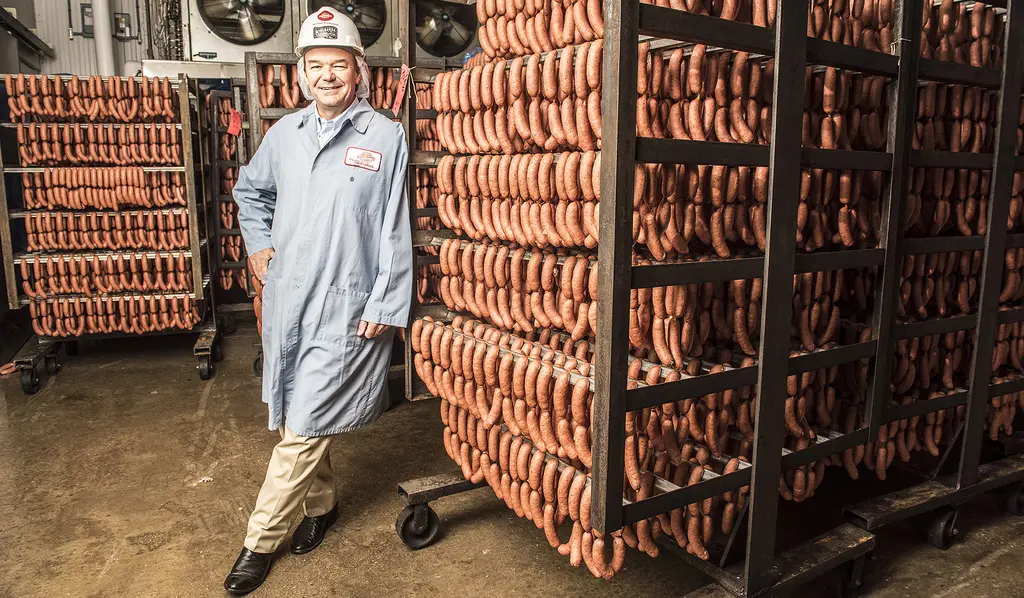 Michael Kiolbassa in a hard hat and blue lab coat stands beside tall racks of smoked sausage