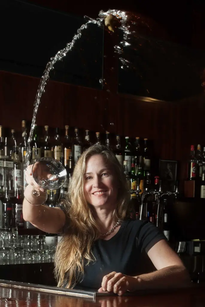 Lisa Owen smiles at a bar as liquid arcs high into the air from the glass in her raised hand