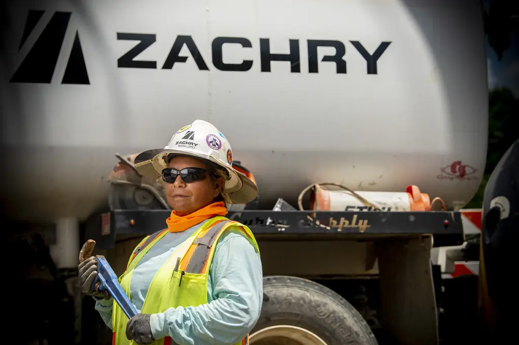 Zachry Construction crew member in a hard hat, sunglasses and safety vest stands in front of a Zachry tank truck