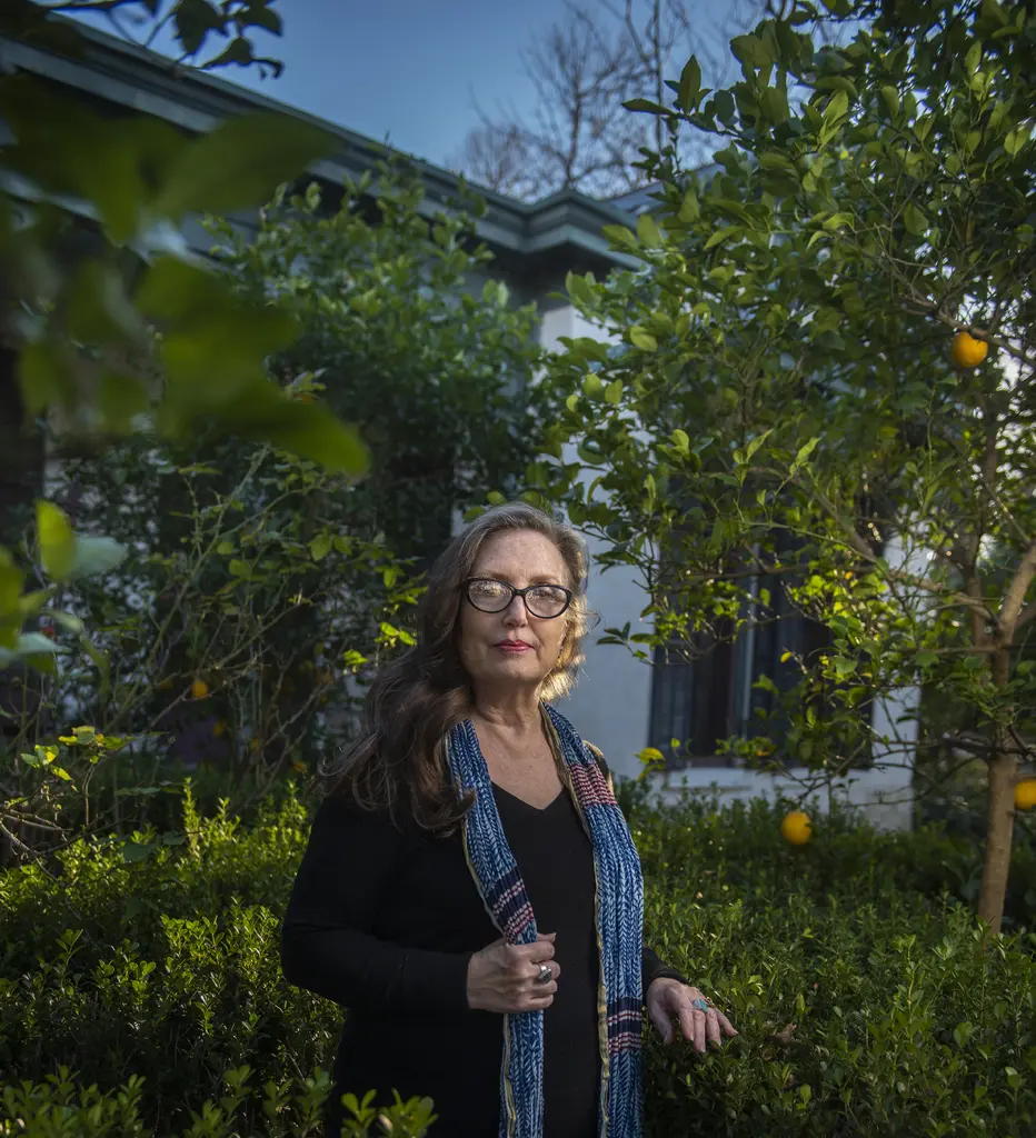 Mary Nethery in glasses and a blue scarf stands among citrus trees outside a house