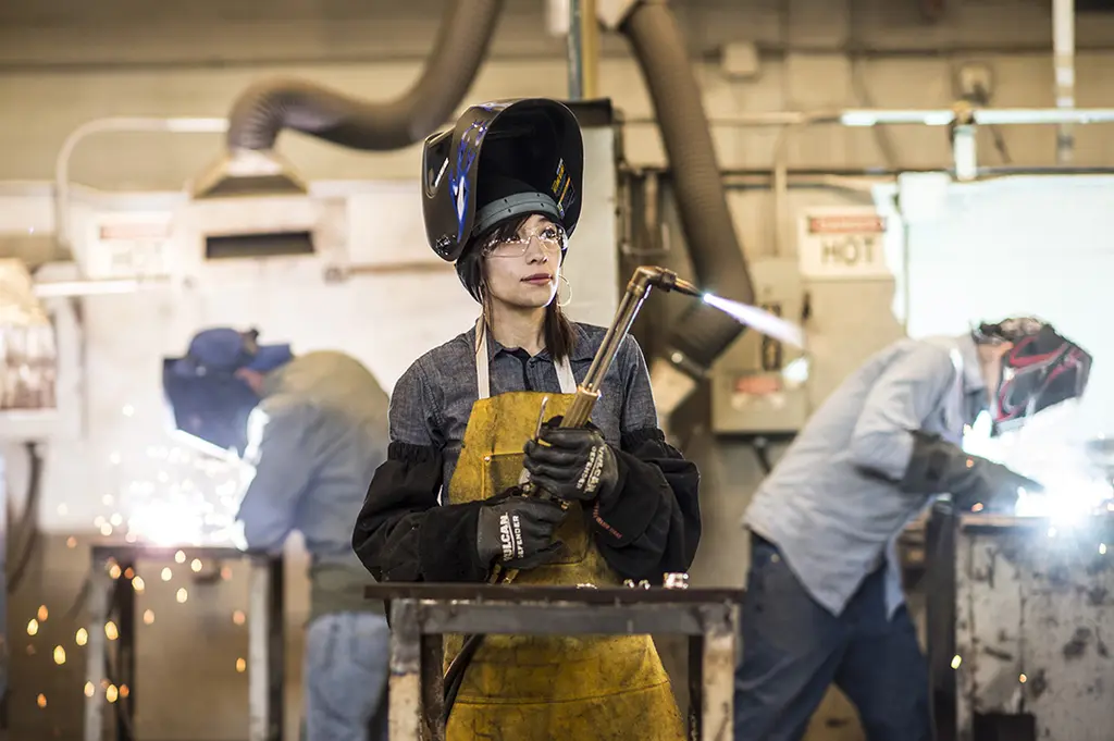 Woman in a flipped-up welding helmet and leather apron holds a torch as two welders work behind her amid sparks