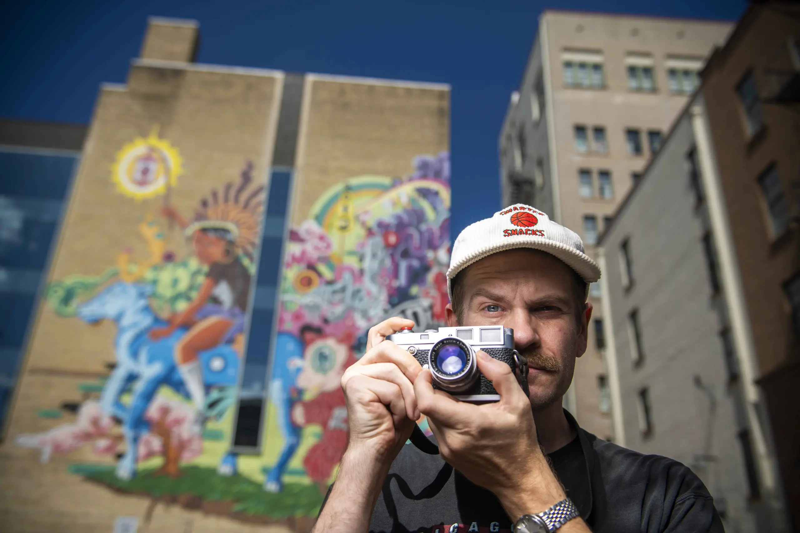 Erik Gustafson in a white cap aiming a film camera in front of a colorful downtown mural