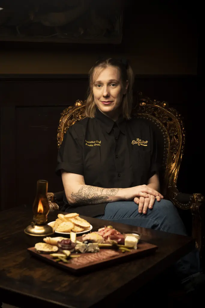Chef Brooke Smith in a black chef's coat sits in an ornate chair behind a charcuterie board