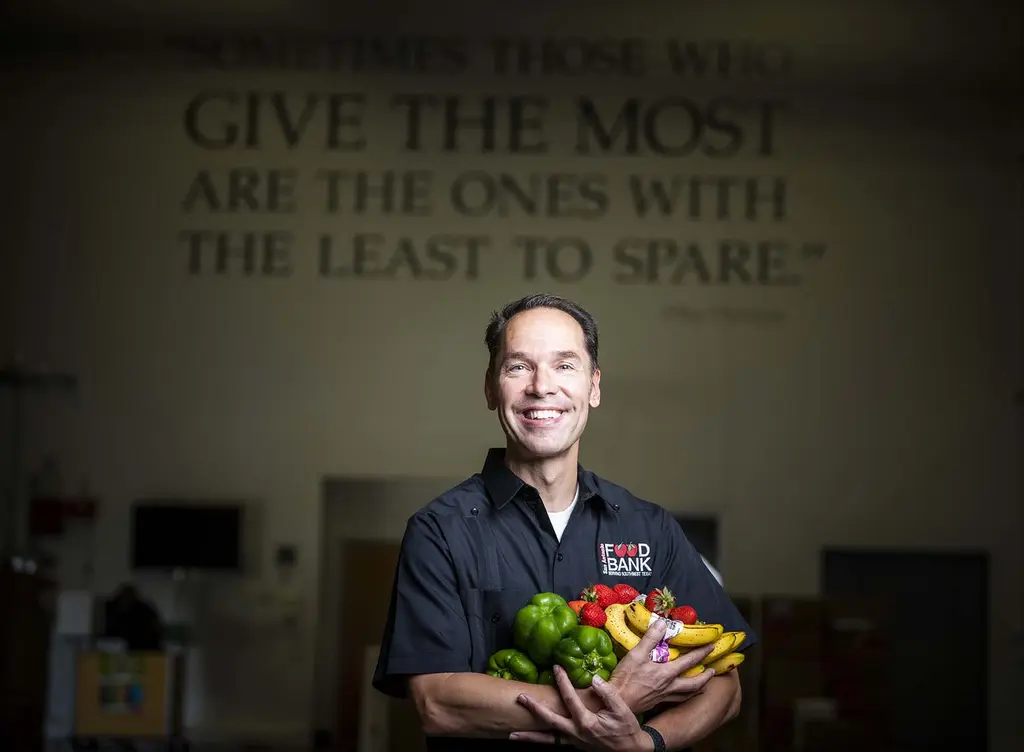 Eric Cooper in a Food Bank shirt holding an armful of fresh produce beneath a painted quote about giving