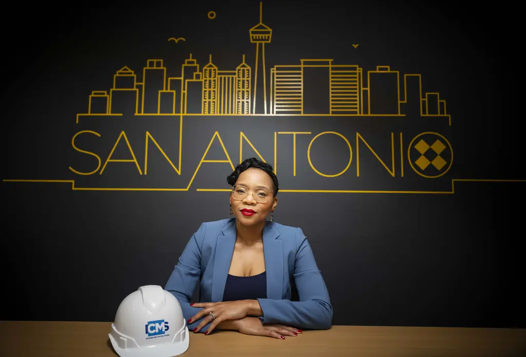 LaShawn Stewart-Baylor in a blue blazer sits at a table with a white hard hat below a gold San Antonio skyline graphic