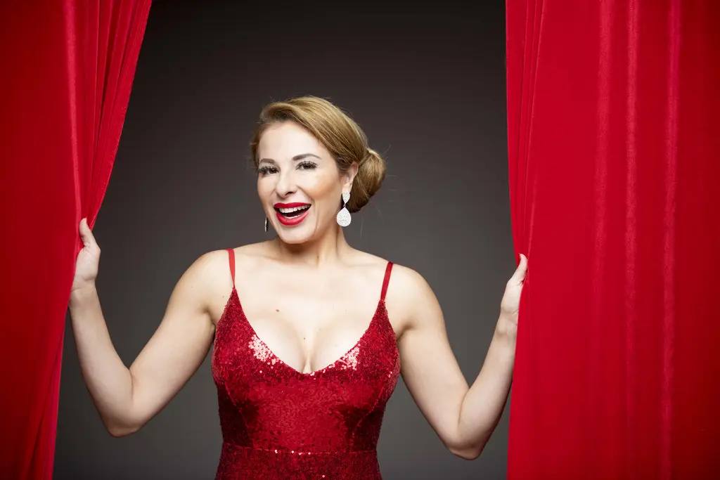 Penny Ayarzagoitia in a red sequined gown smiles as she holds open a pair of red stage curtains
