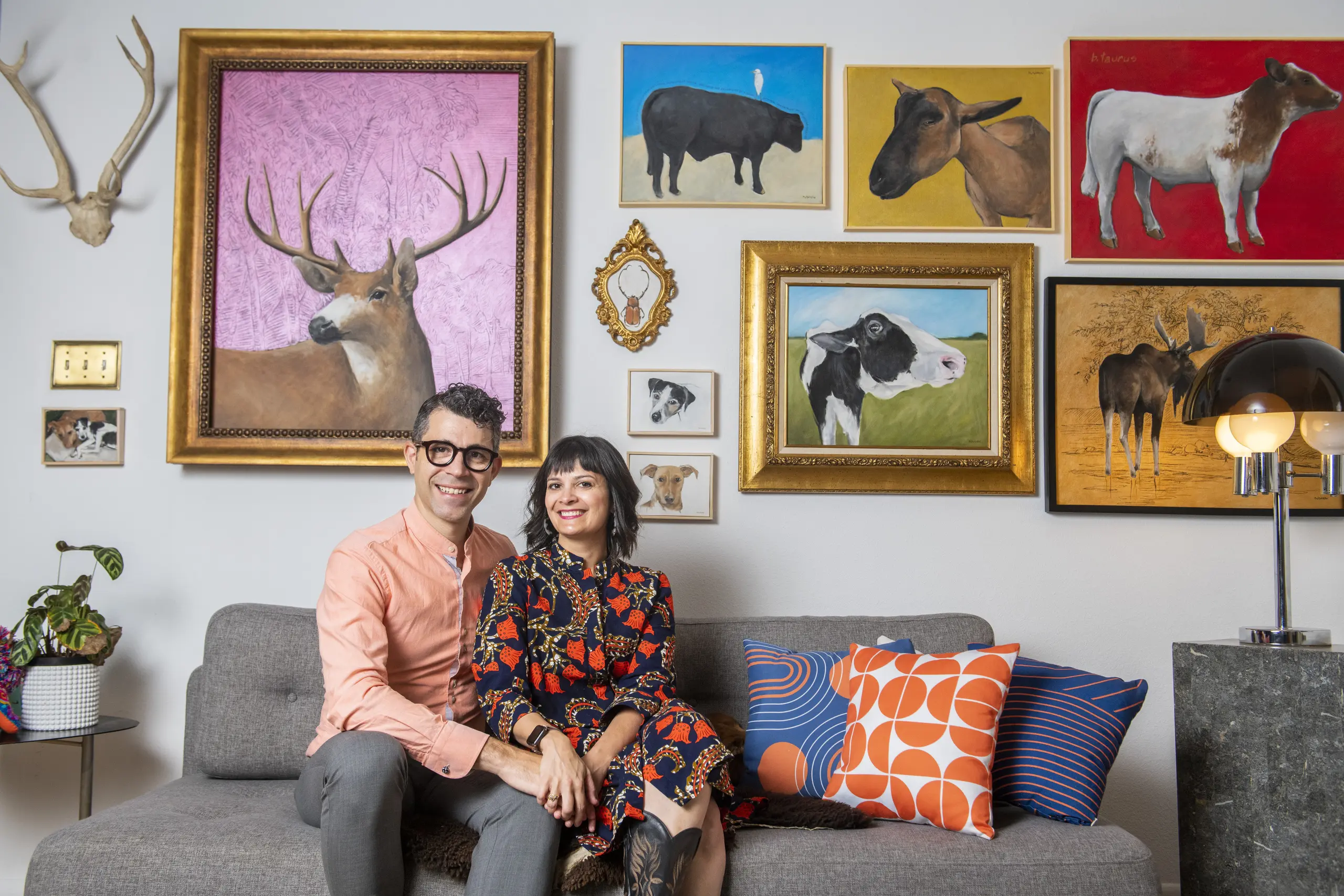 Marisa Bushman and Ignacio Gallego sit on a gray sofa beneath a wall of framed animal paintings