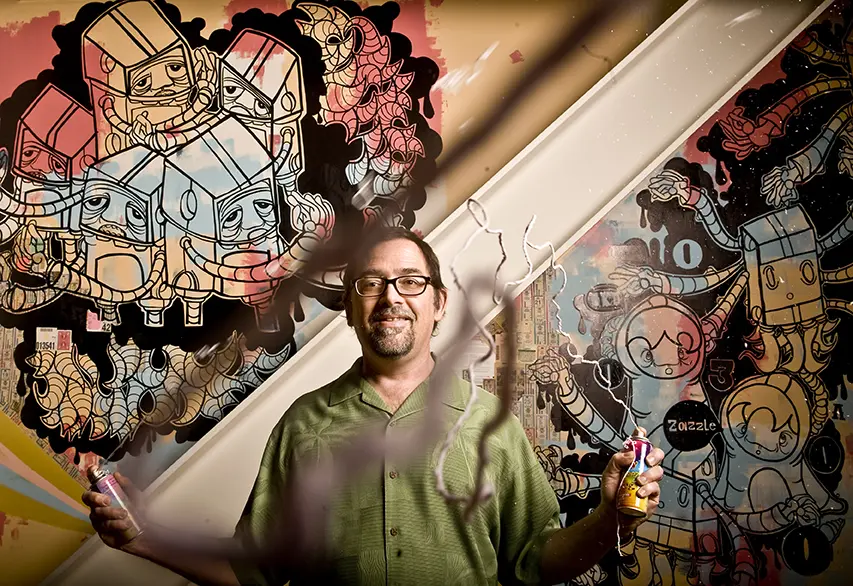 Young Harvill in glasses and a green shirt holds spray cans as string streams across a colorful mural