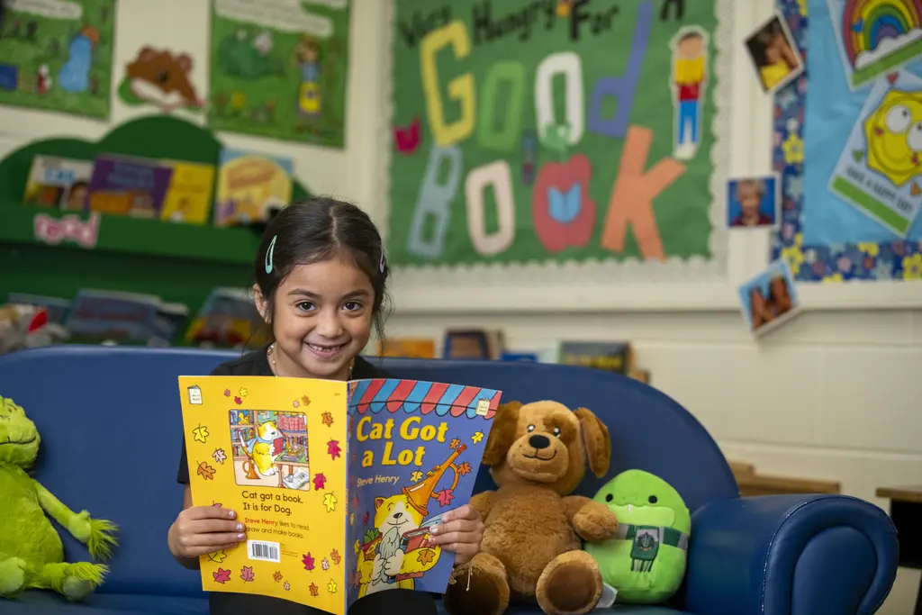 Young girl smiling over the book Cat Got a Lot on a classroom couch beside stuffed animals