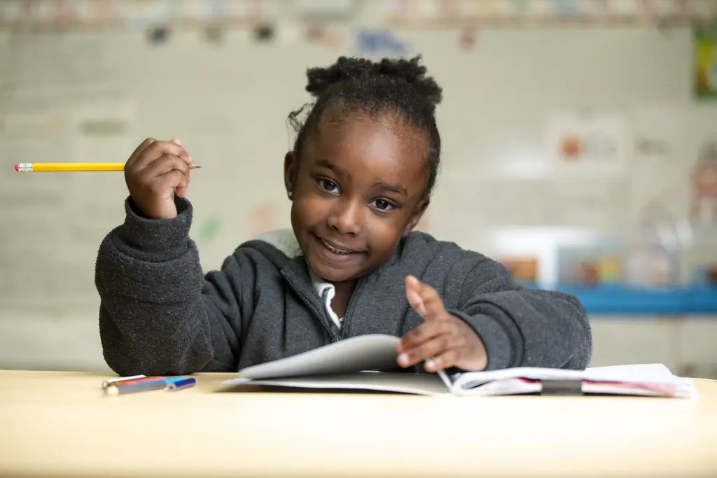Young girl in a gray hoodie smiling at her desk with a pencil raised over an open notebook