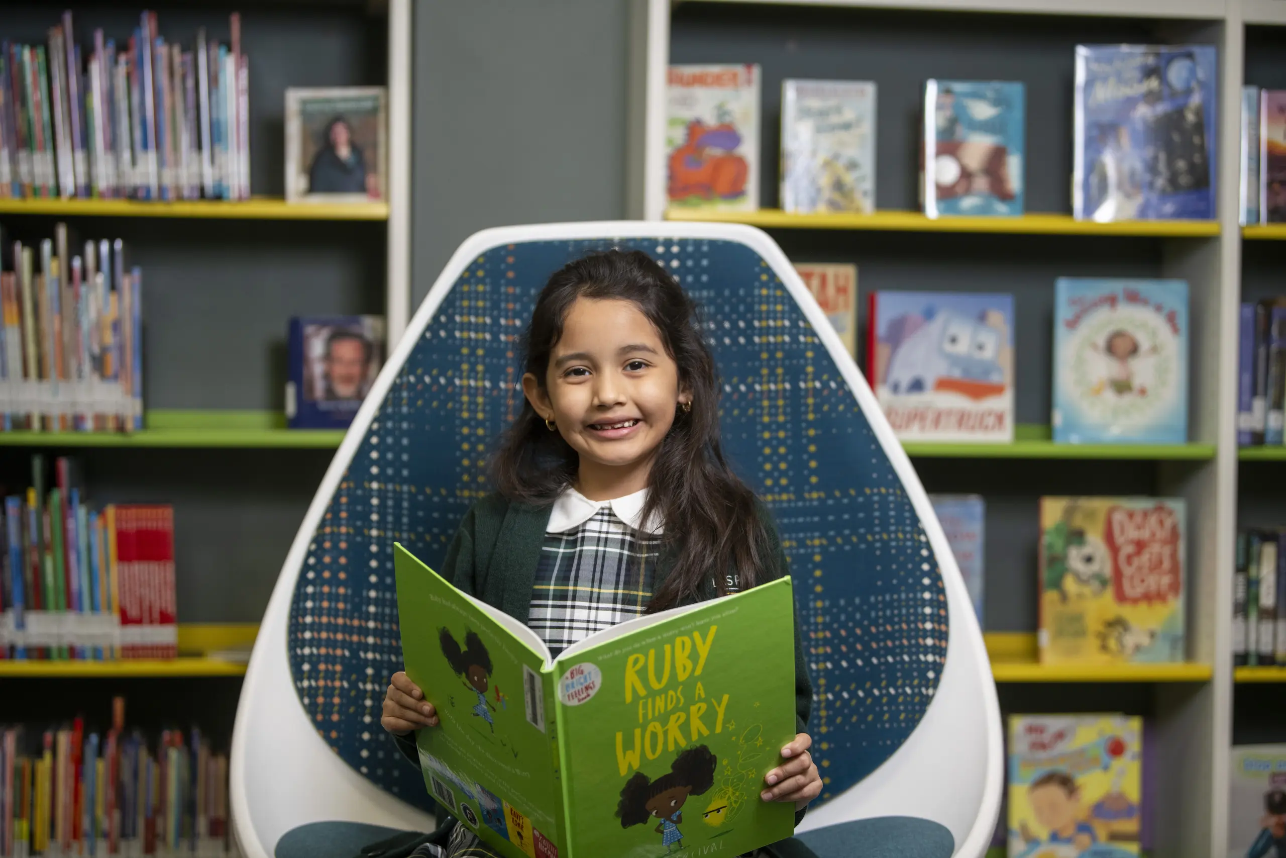 Young girl in a plaid school uniform holding the book Ruby Finds a Worry in a library chair