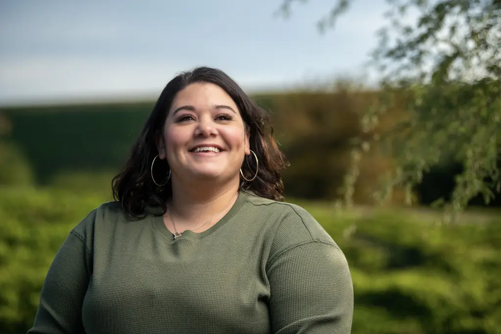 Geneva Knott, in an olive green waffle-knit top and silver hoop earrings, smiles outdoors with green trees and shrubs softly blurred behind her.