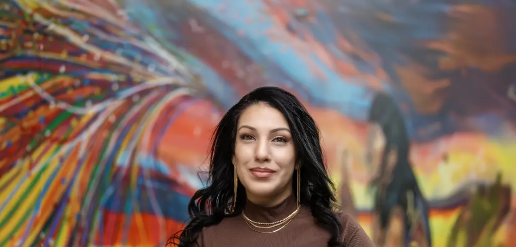 Josie Perez, in a brown top, stands in front of a colorful painted mural.