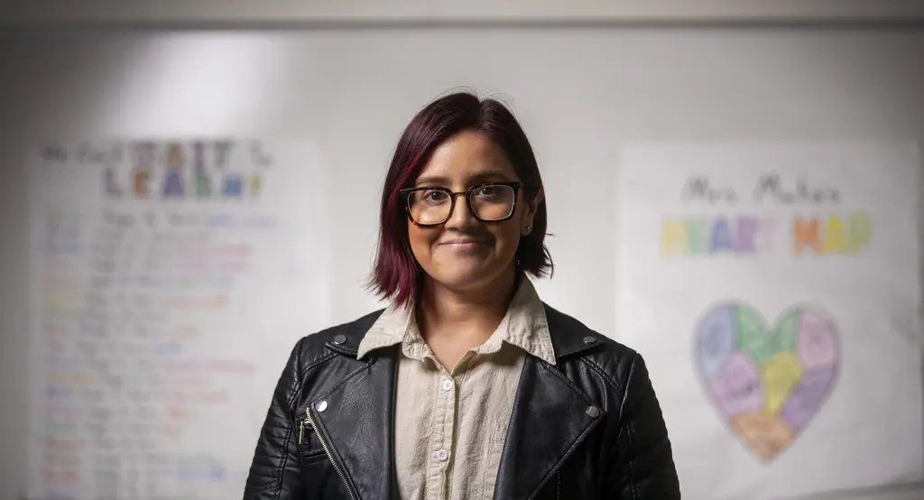 Teacher Ms. Mata in glasses and a black leather jacket in front of classroom posters