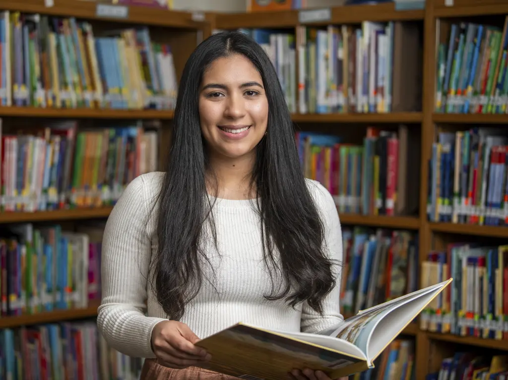Yesenia Villafranco in a white sweater holding an open book in front of library shelves