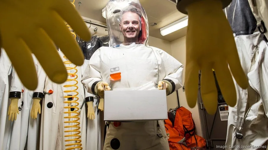 Scientist Robert Davey, in a protective suit, smiles while holding a white box, framed by yellow lab gloves.