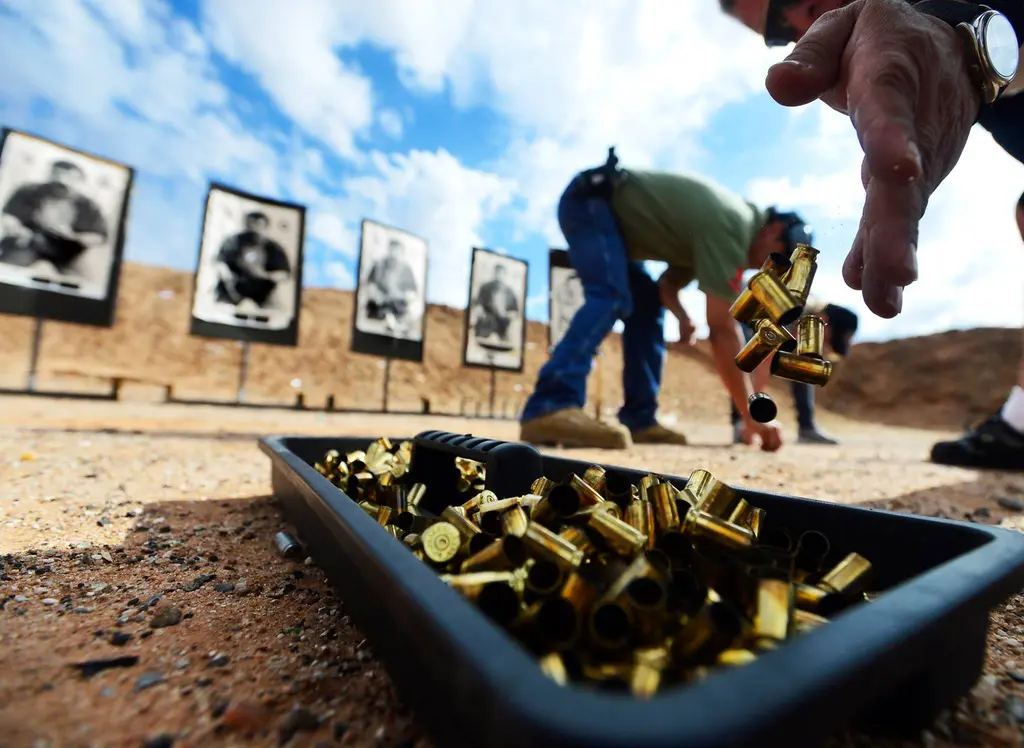 A hand drops spent brass shell casings into a black tray in the dirt as a shooter bends to pick up more in front of a row of paper targets.