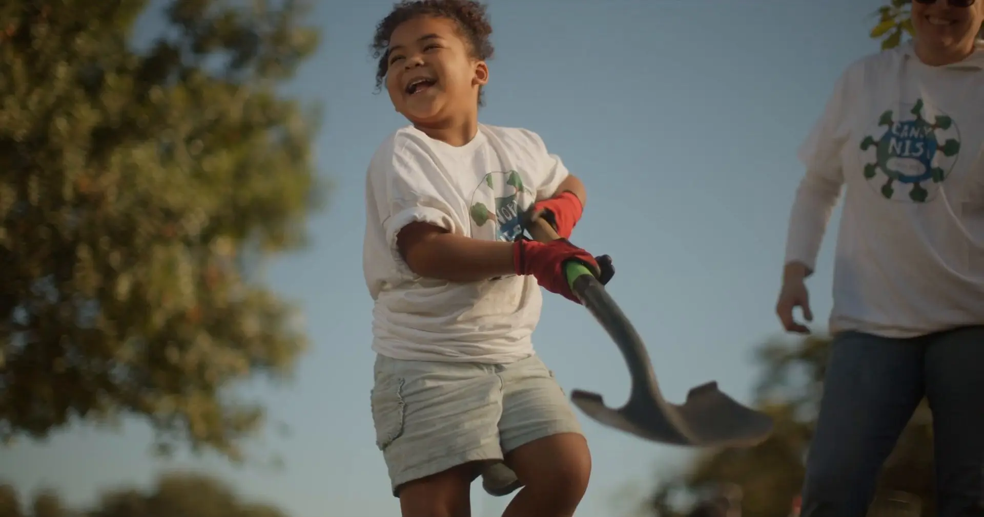 A laughing girl in red work gloves and a white T-shirt swings a shovel under a blue sky as a smiling adult in a white hoodie stands nearby.