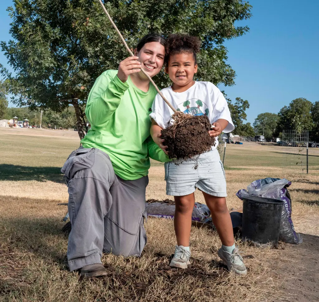 Desiree Mendoza, vestida de verde, se arrodilla y sujeta el tallo de un arbolito junto a su hija, que sonríe y sostiene las raíces cubiertas de tierra en un parque.
