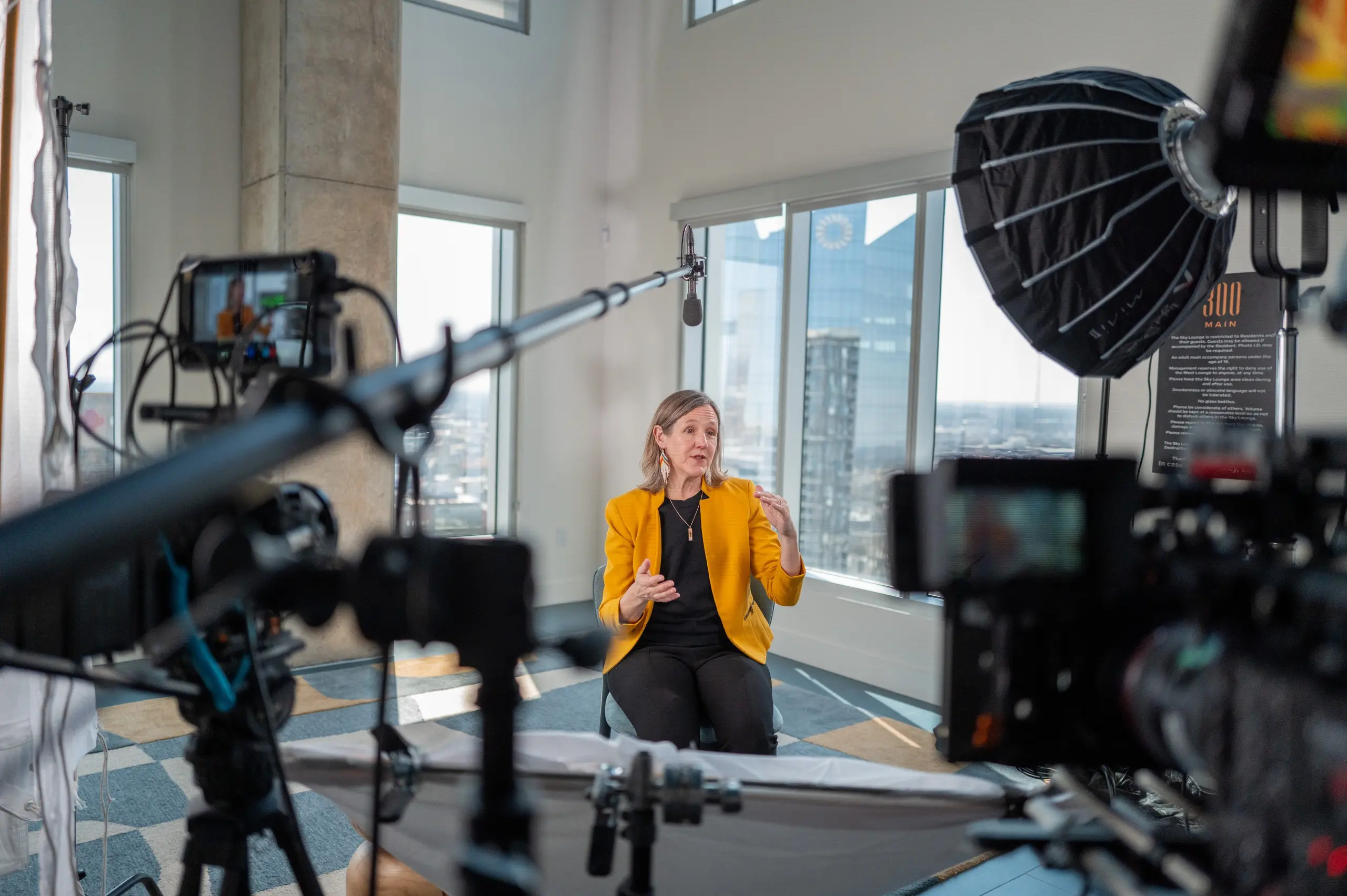 Jeanne Russell of CAST Schools, in a mustard-yellow blazer, gestures during an interview by tall windows, framed by a boom mic, a large softbox and two cinema cameras.