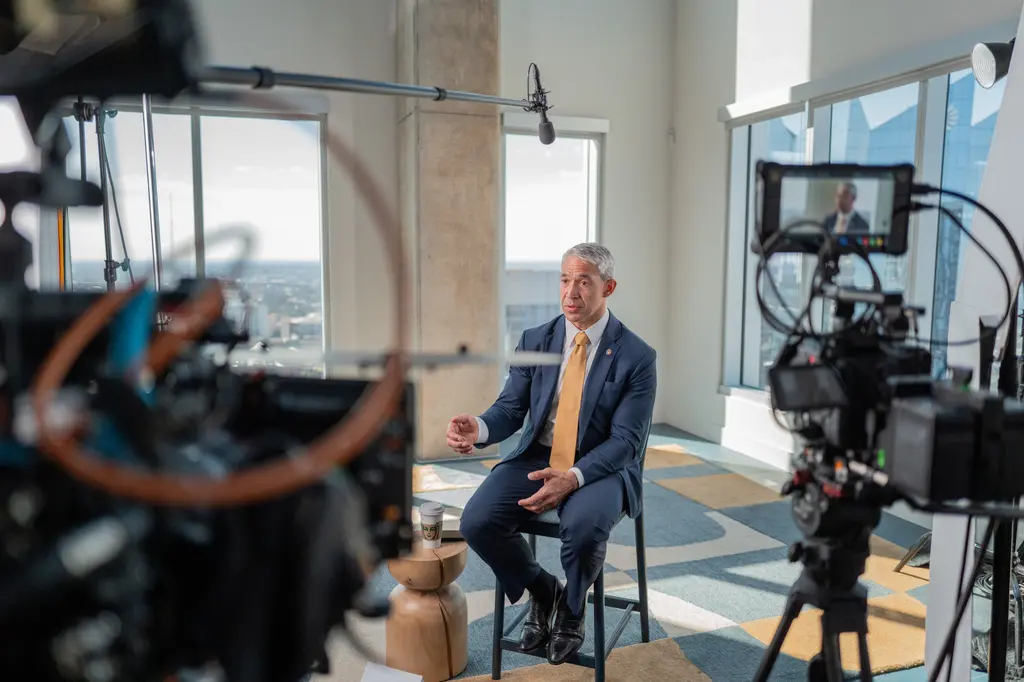 El exalcalde de San Antonio Ron Nirenberg, de traje azul marino y corbata dorada, habla con las manos durante una entrevista junto a unos ventanales altos, mientras dos cámaras de cine y un micrófono boom graban.