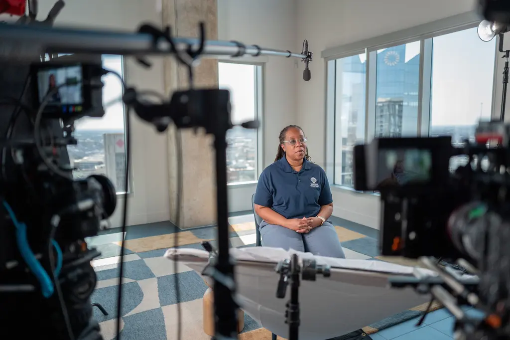 Sonya Bryant, in a navy polo, sits for an interview by tall windows overlooking downtown, framed by two cinema cameras and a boom mic.