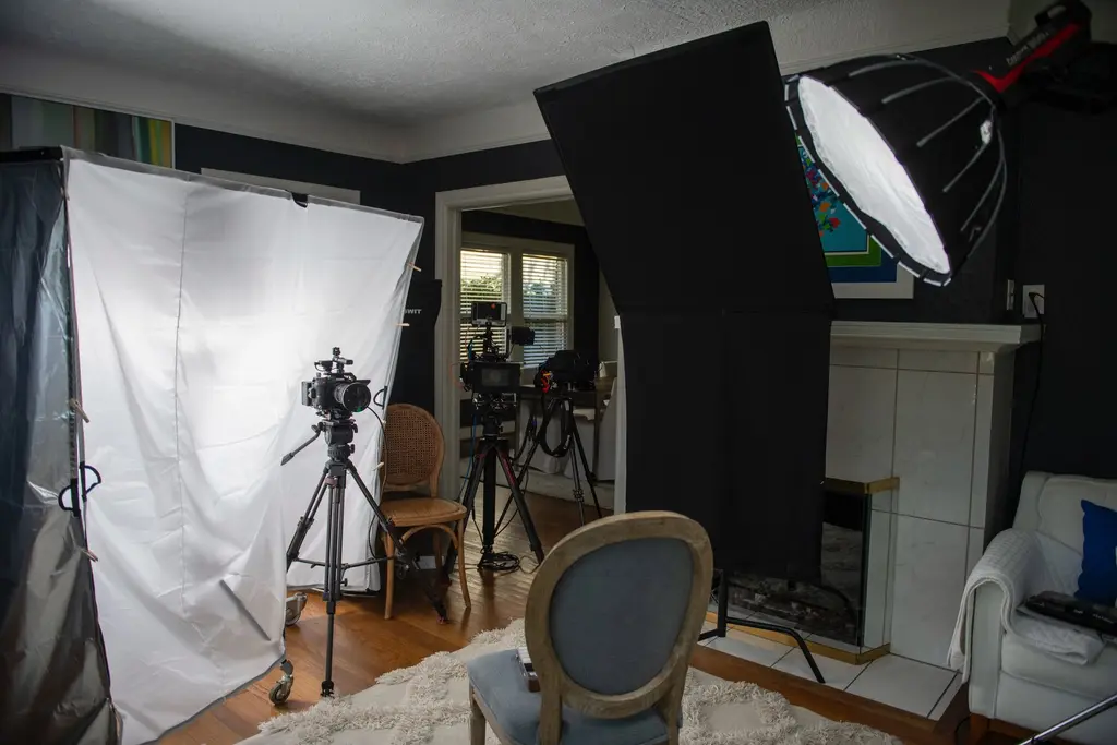 Cameras on tripods face an empty gray chair by a white fireplace in a dark-walled living room, lit by a large softbox and a white diffusion panel.