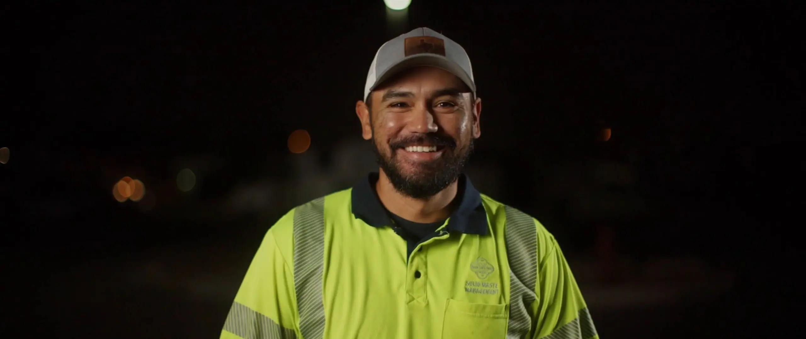 A smiling, bearded man in a light cap and a bright yellow Solid Waste Management safety shirt stands outside at night.