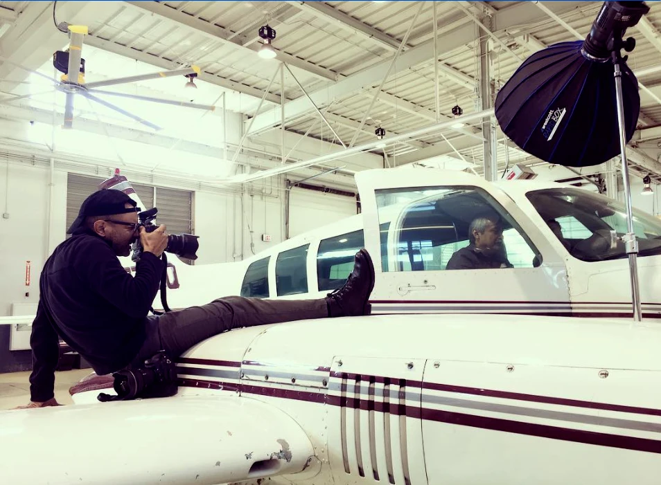 Carlos Javier Sanchez lies on the wing of a small plane in a St. Philip’s College hangar, photographing an aviation instructor in the cockpit under a softbox.