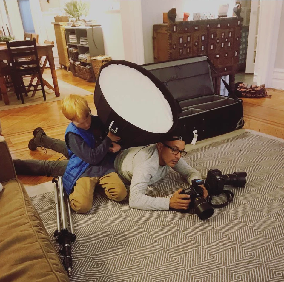 Carlos Javier Sanchez lies on a living-room rug with his cameras while a young boy helps hold a large softbox, on assignment for Petco Love.