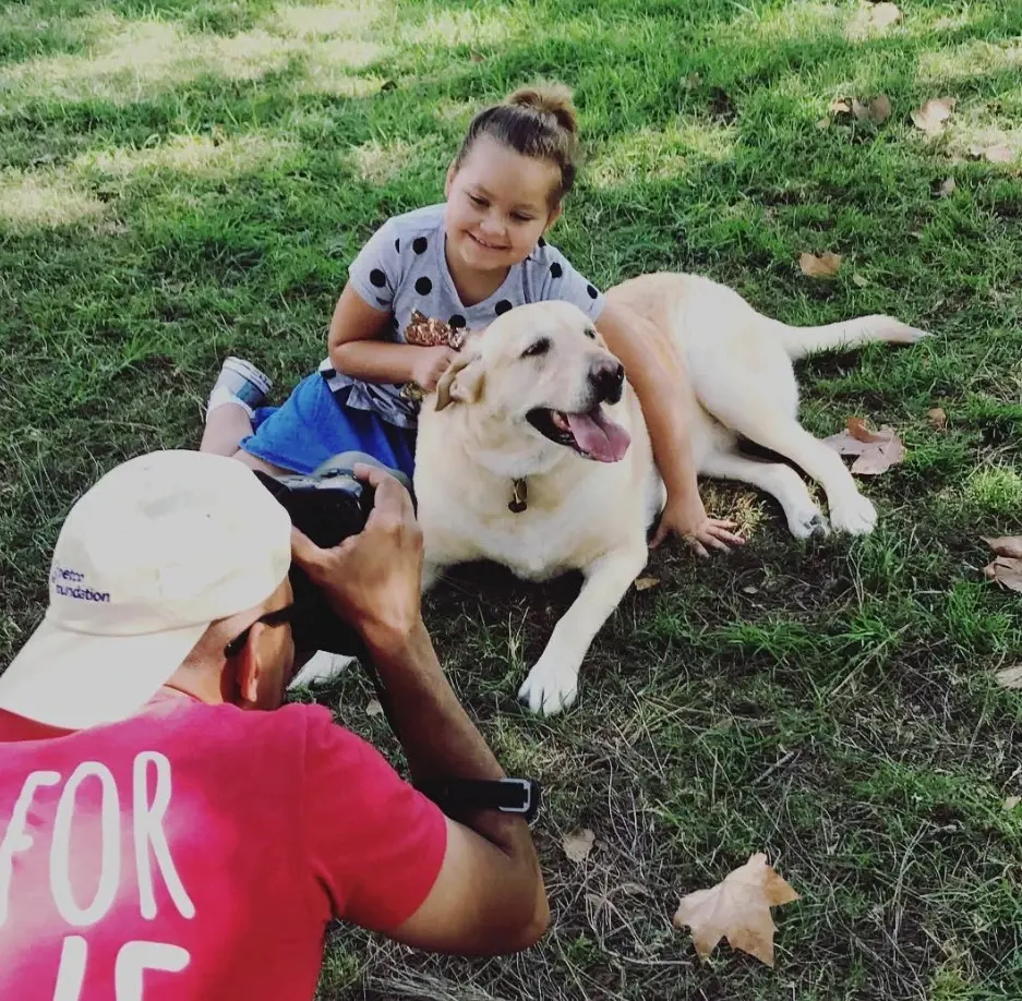 Up close, Carlos Javier Sanchez, in a pink All For Love shirt, photographs a smiling girl and her yellow Labrador lying in the grass, on assignment for Petco Love.