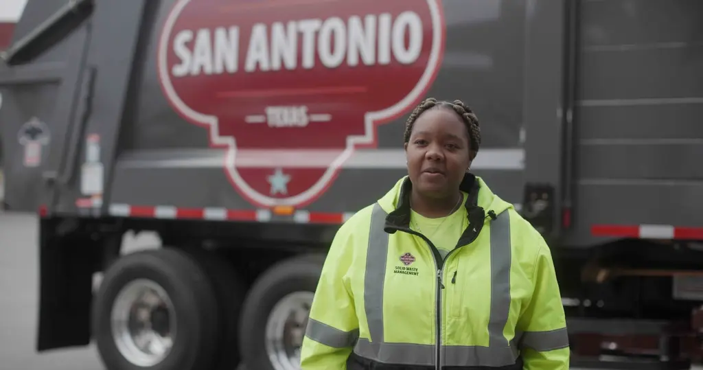 A woman in a high-visibility jacket stands in front of a garbage truck with the San Antonio logo.