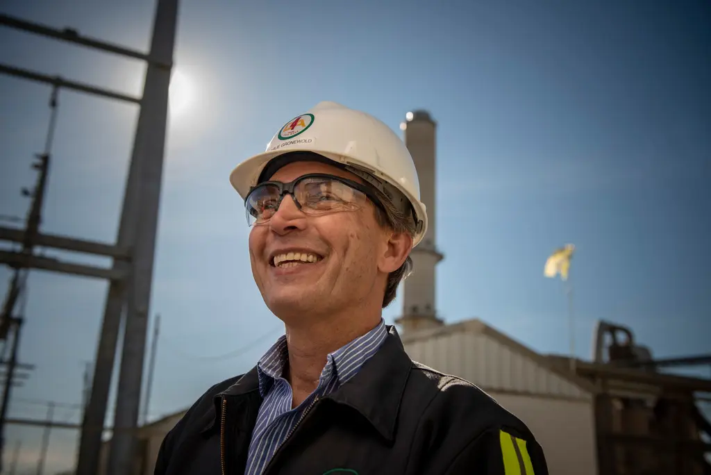 Charles Gronewold in a white hard hat and safety glasses smiles near an industrial stack