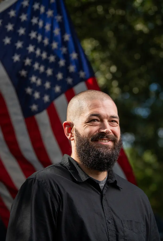 Charlie Wright in a black shirt smiles in front of an American flag