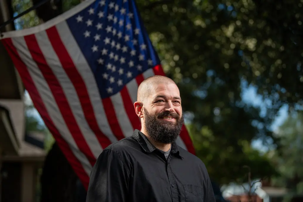 Charlie Wright in a black shirt smiles in front of an American flag and shade trees