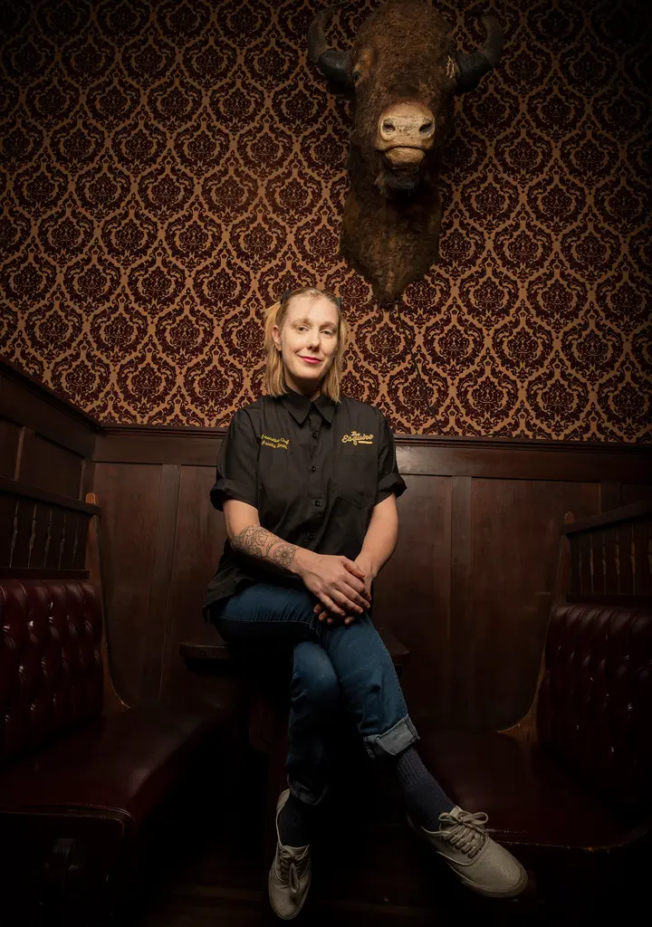 Chef Brooke Smith sits with her legs crossed between two leather booths, a bison head mounted on the wall above her.