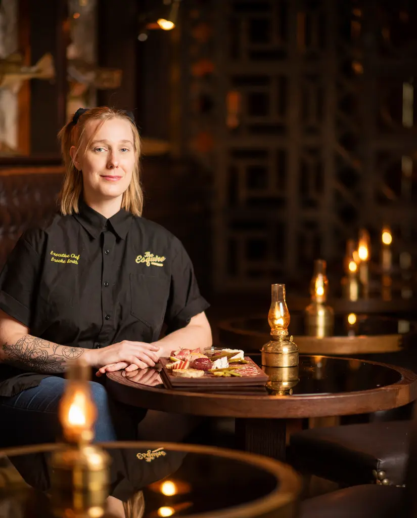 Chef Brooke Smith sits at a dark round table with a charcuterie board and a lit oil lamp.