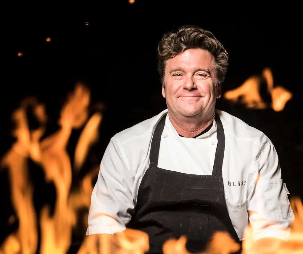 Chef Mark Bliss, in a white chef coat and black Bliss apron, smiles behind leaping flames.