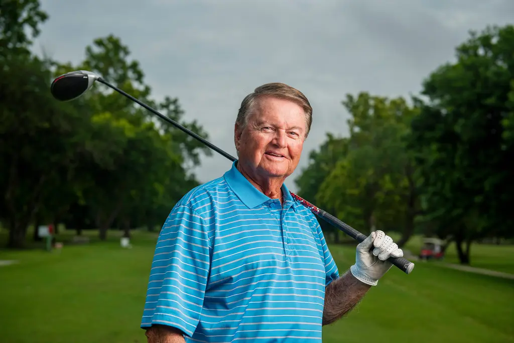 A smiling golfer in a striped blue polo rests a driver on his shoulder on a tree-lined fairway.
