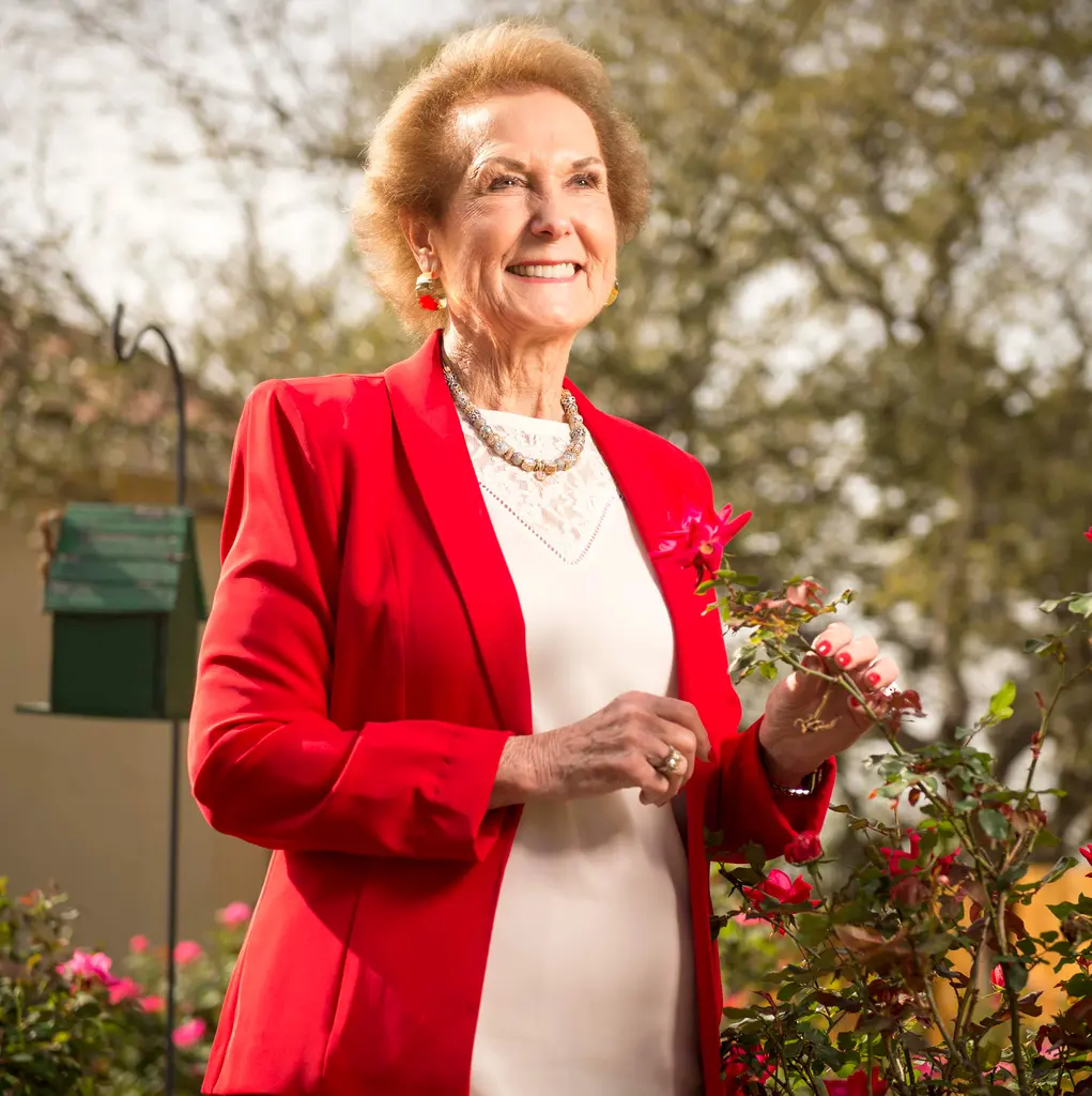D'Ann Harper in a red jacket smiles as she touches a rose in a garden