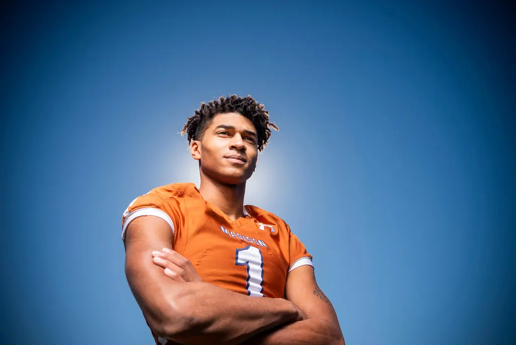 A young quarterback in an orange Madison No. 1 jersey stands with his arms crossed against a deep blue sky.