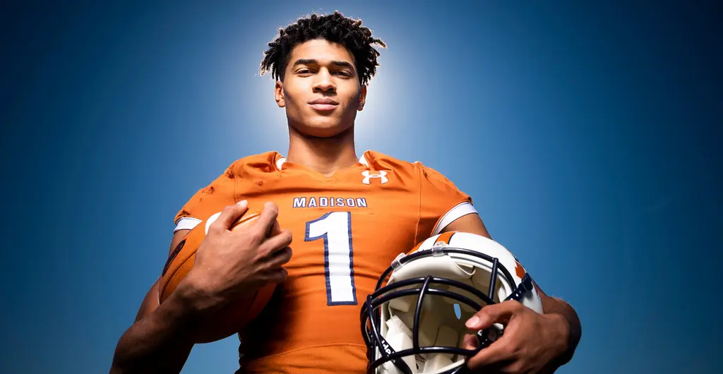 A young quarterback in an orange Madison No. 1 jersey holds a football and a white helmet, photographed from below against a blue sky.