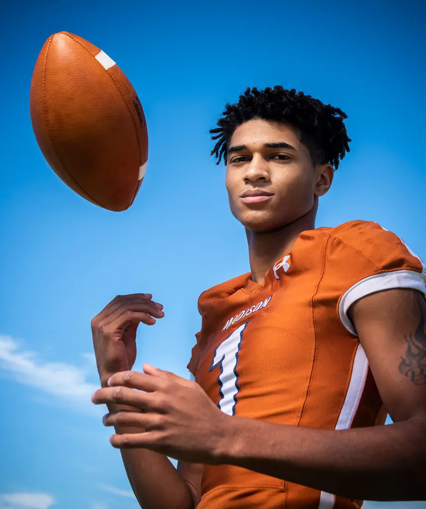 A young quarterback in an orange Madison No. 1 jersey looks down at the camera as a football hangs in the air beside him.