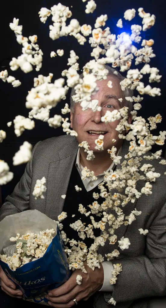 Dennis Noll in a houndstooth jacket smiles through a burst of popcorn flying from a bag