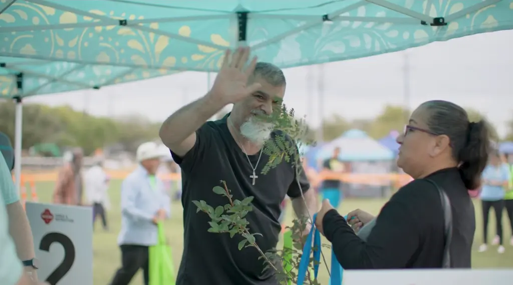 A man with a gray beard holds a young tree and waves at a Parks and Recreation giveaway tent as a woman beside him holds a blue tote bag.