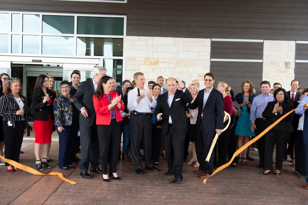 A crowd in business attire claps and cheers in front of the hotel's glass doors as a man holding oversized gold scissors smiles and lengths of orange ribbon fall to the pavement.