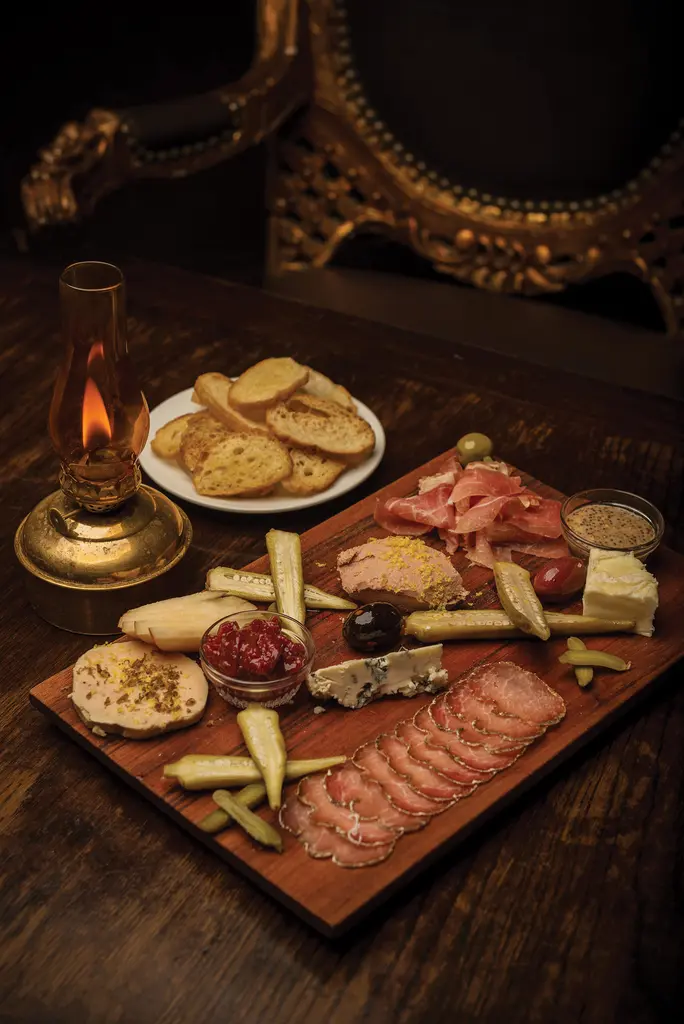 A charcuterie board and a plate of toasted bread beside a lit brass oil lamp on a dark wooden table.