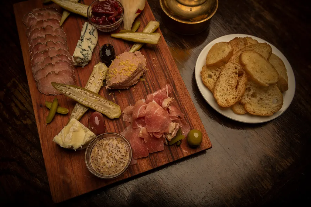 An overhead view of a wooden charcuterie board with cured meats, blue cheese, pâté, pickled okra and olives beside a plate of toasted bread.