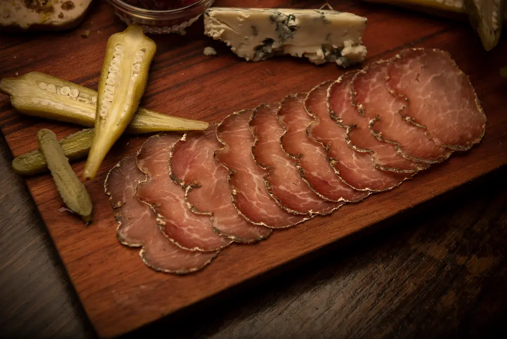 Thin slices of cured meat fanned across a wooden board beside pickled okra and blue cheese.